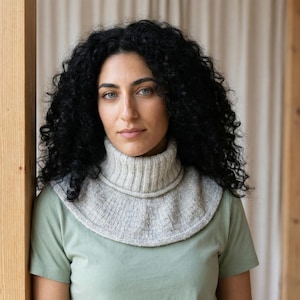 May include: A light gray knitted cowl with a ribbed turtleneck and a flared lower edge. The cowl is worn over a light green t-shirt. The person in the image has dark curly hair and is leaning against a wooden structure.