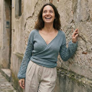 May include: A light blue wrap sweater with textured sleeves and a V-neck. The sweater is paired with beige pleated pants. The person is smiling and leaning against a textured stone wall.