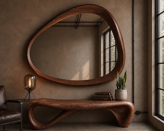 May include: A large, organic-shaped wall mirror with a thick, dark wood frame. Below it sits a matching, fluidly curved wooden console table. A small glass lamp with a visible filament bulb and a potted snake plant rest on the table surface.
