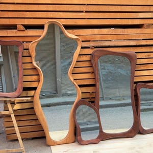 May include: A collection of five mirrors with irregular, organic-shaped wooden frames in varying shades of brown. The mirrors feature smooth, curved edges and reflective glass surfaces, displayed together in a group with different sizes and shapes.