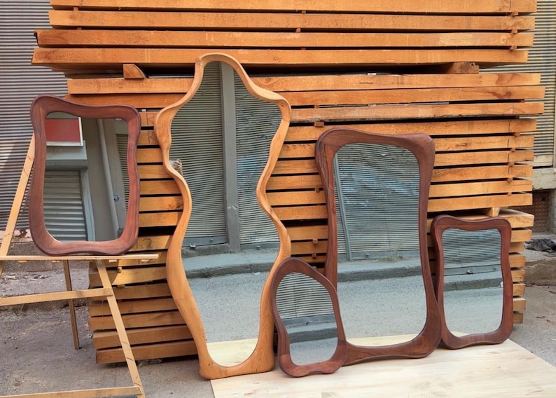 May include: A collection of five mirrors with irregular, organic-shaped wooden frames in varying shades of brown. The mirrors feature smooth, curved edges and reflective glass surfaces, displayed together in a group with different sizes and shapes.
