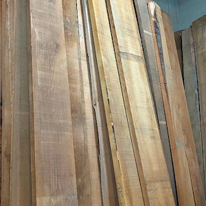May include: A stack of long, unfinished wooden planks standing vertically. The boards feature natural light to medium brown wood grain patterns, with some edges showing raw bark. One board has the handwritten text ADET-A1 visible on its surface.