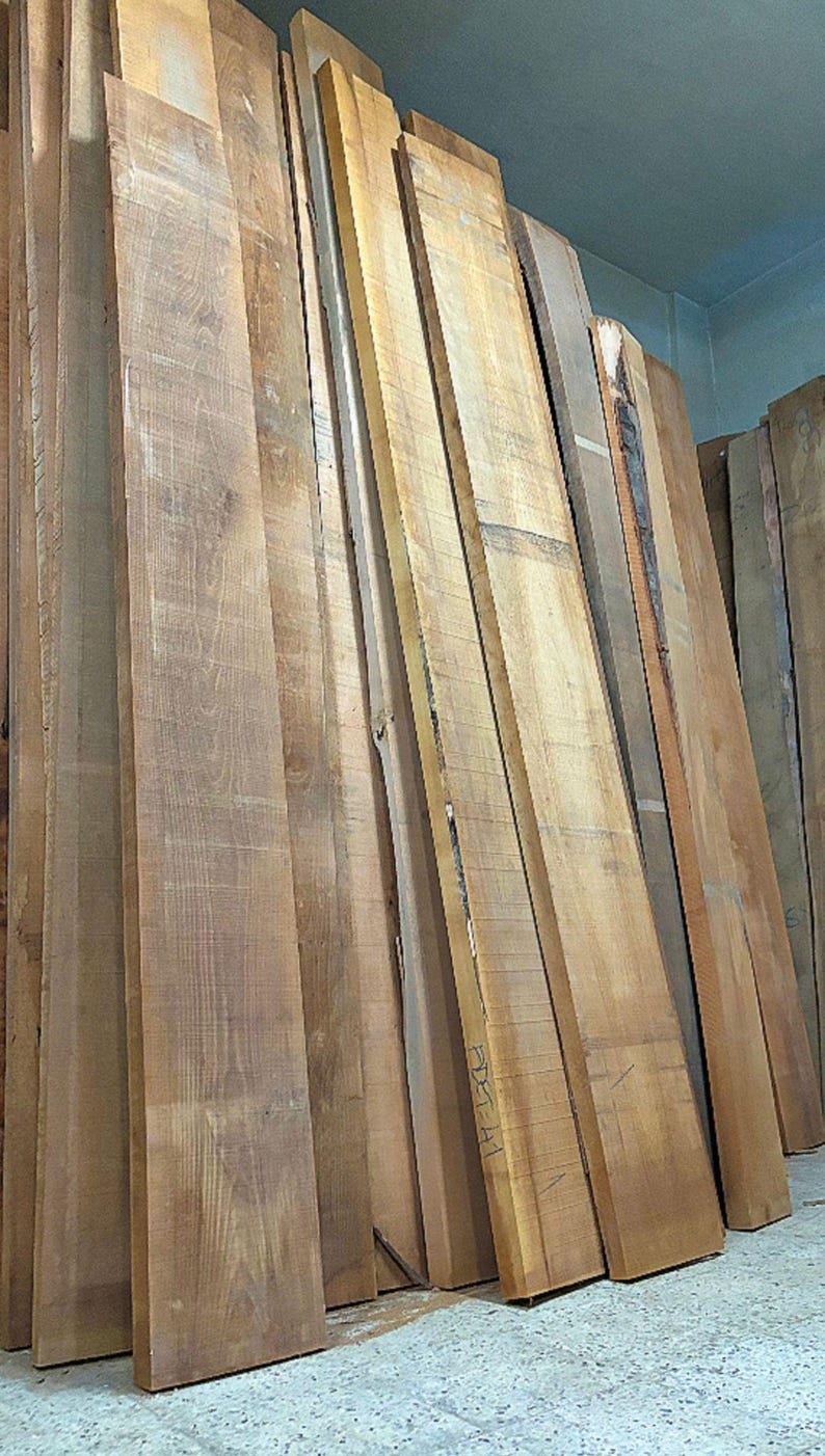May include: A stack of long, unfinished wooden planks standing vertically. The boards feature natural light to medium brown wood grain patterns, with some edges showing raw bark. One board has the handwritten text ADET-A1 visible on its surface.