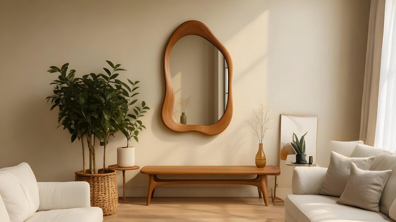 Puede incluir: Un espejo de pared con forma org&aacute;nica irregular y un marco de madera lisa de color marr&oacute;n claro. El espejo est&aacute; montado en una pared beige lisa sobre un banco de madera largo y bajo con bordes redondeados y cuatro patas, creando un conjunto.