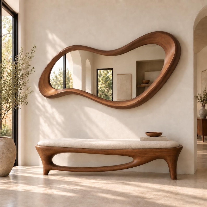 May include: A large, wall-mounted mirror with an irregular, organic wooden frame featuring a smooth, flowing shape. Below the mirror sits a long, low wooden bench with a cream-coloured upholstered seat and a hollow, curved base design.
