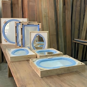 May include: Several mirrors of varying shapes and sizes are secured within light-colored wooden shipping crates. Each mirror is protected by blue foam padding along the edges and white protective sheets, arranged on a large wooden workbench.