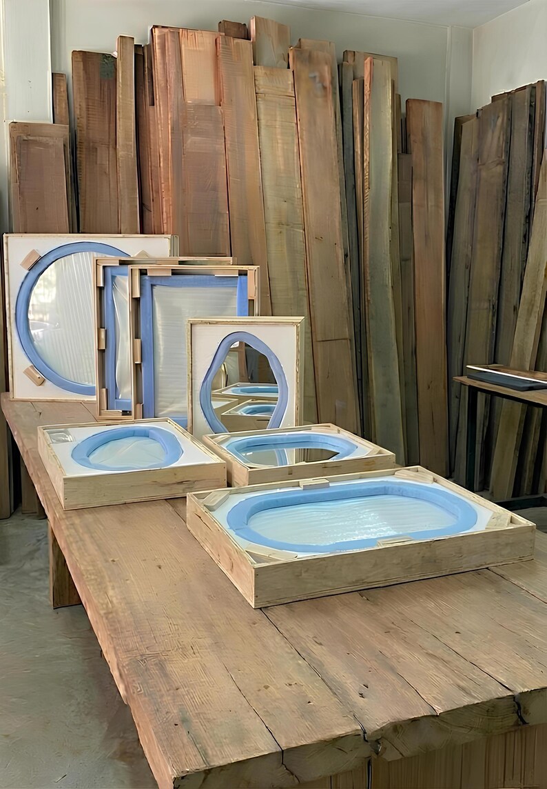 May include: Several mirrors of varying shapes and sizes are secured within light-colored wooden shipping crates. Each mirror is protected by blue foam padding along the edges and white protective sheets, arranged on a large wooden workbench.