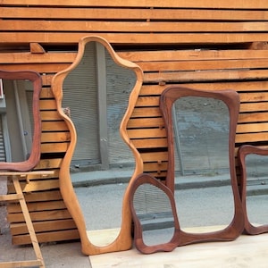 May include: A collection of four wooden mirrors with irregular, organic shapes and smooth, rounded frames. The mirrors vary in size and wood tone, ranging from light to dark brown, and are displayed together in a group with reflective glass surfaces.