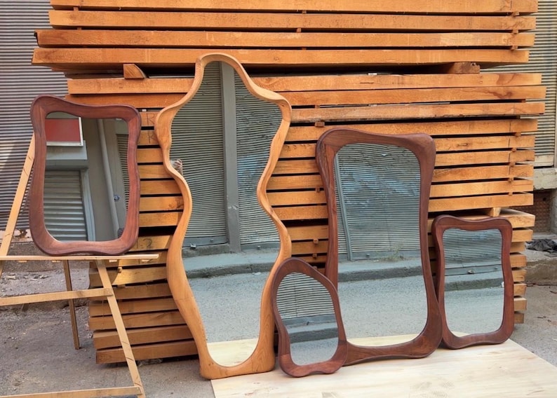 May include: A collection of four wooden mirrors with irregular, organic shapes and smooth, rounded frames. The mirrors vary in size and wood tone, ranging from light to dark brown, and are displayed together in a group with reflective glass surfaces.