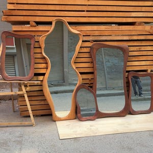 Puede incluir: Una colecci&oacute;n de cuatro espejos de madera con formas org&aacute;nicas irregulares y marcos curvos y lisos. Los espejos var&iacute;an en tama&ntilde;o y tono de madera, desde marr&oacute;n claro a oscuro, y se muestran apoyados contra una pila de madera.