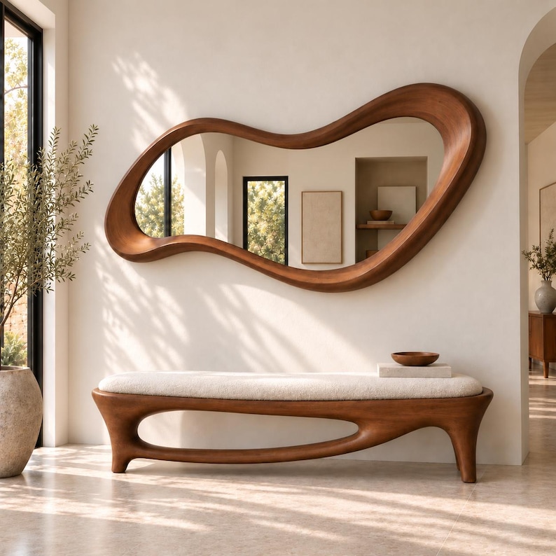 May include: A large, horizontal wall mirror with an irregular, organic wooden frame featuring a smooth, warm brown finish. Below the mirror sits a long bench with a thick, cream-coloured upholstered seat and a curved, solid wood base.