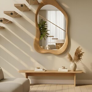 Puede incluir: Un espejo de pared con forma org&aacute;nica irregular y marco de madera clara. El espejo est&aacute; montado en una pared beige sobre un banco de madera largo y rectangular. El reflejo muestra una planta verde en maceta y parte de una escalera.