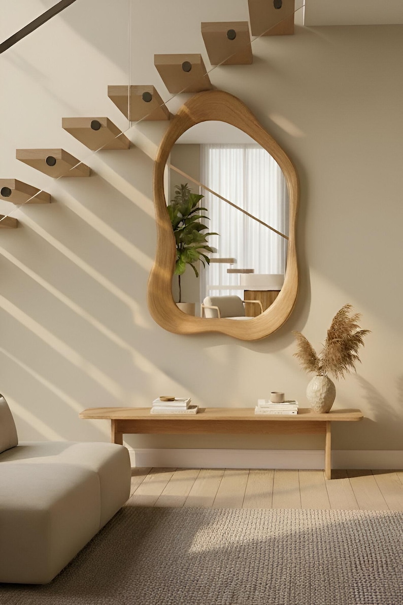 Puede incluir: Un espejo de pared con forma org&aacute;nica irregular y marco de madera clara. El espejo est&aacute; montado en una pared beige sobre un banco de madera largo y rectangular. El reflejo muestra una planta verde en maceta y parte de una escalera.