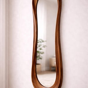 May include: A tall, full-length wall mirror featuring a smooth, organic, wavy wooden frame with a rich brown finish. The mirror has an elongated, asymmetrical shape that curves inward along the sides, creating a fluid and continuous wooden border.