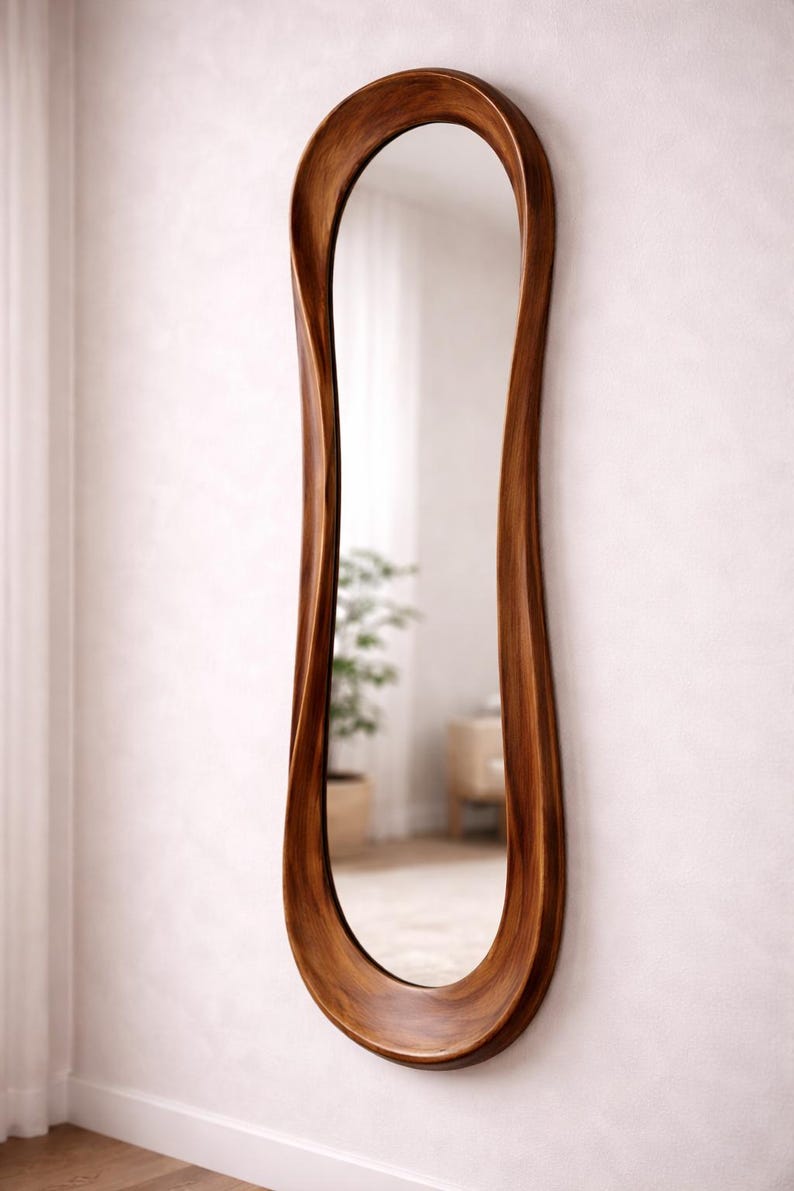 May include: A tall, full-length wall mirror featuring a smooth, organic, wavy wooden frame with a rich brown finish. The mirror has an elongated, asymmetrical shape that curves inward along the sides, creating a fluid and continuous wooden border.