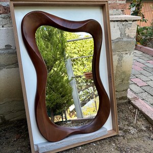 May include: A wall mirror featuring a dark brown, smooth, organic, wavy wooden frame. The mirror is mounted within a rectangular, light-coloured wooden box frame that provides a contrasting border. The reflective surface shows a garden scene.