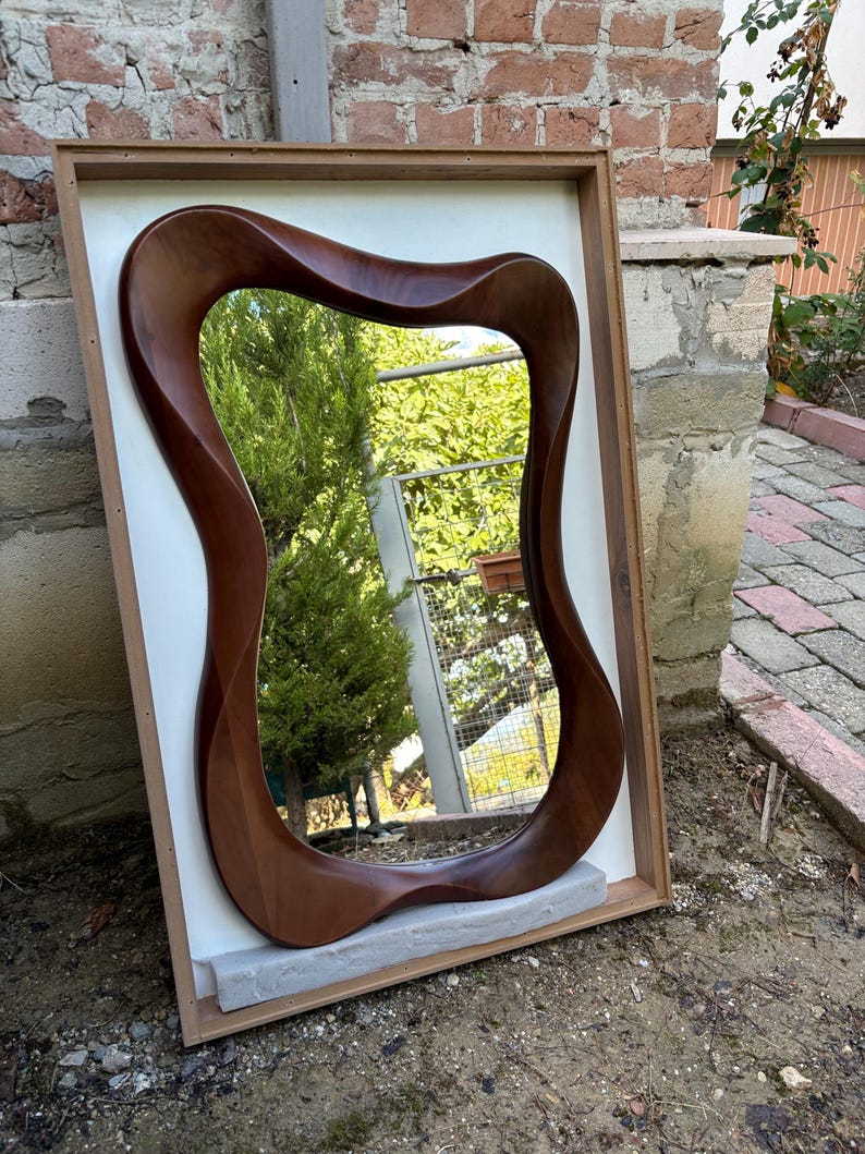 May include: A wall mirror featuring a dark brown, smooth, organic, wavy wooden frame. The mirror is mounted within a rectangular, light-coloured wooden box frame that provides a contrasting border. The reflective surface shows a garden scene.