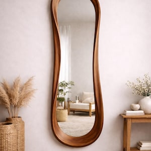 May include: A full-length wall mirror featuring a smooth, organic, wavy wooden frame with a rich brown finish. The mirror has an elongated, asymmetrical shape that tapers slightly toward the top and bottom, reflecting a bright interior space.