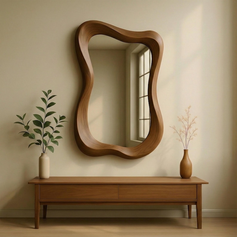 May include: A wall-mounted mirror with a thick, wavy, organic-shaped wooden frame. The mirror is positioned above a long, rectangular wooden console table with two drawers. A light-colored vase with green leaves sits on the left, and a brown vase with thin, dried branches sits on the right.