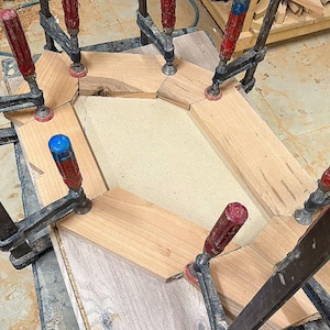 May include: A wooden frame assembly held together by multiple metal clamps with red and blue handles. The frame consists of several light-colored wood segments arranged in a hexagonal shape around a central beige panel, showing raw edges.