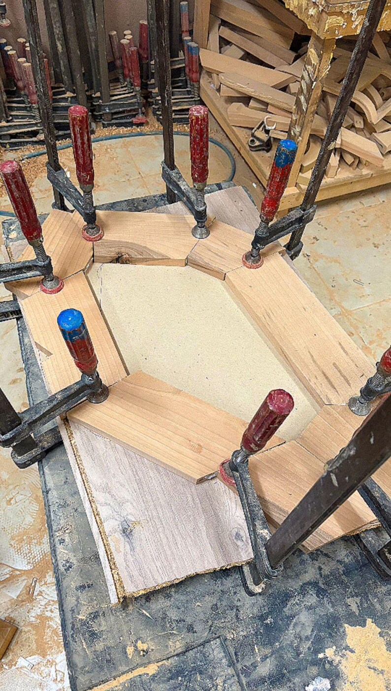 May include: A wooden frame assembly held together by multiple metal clamps with red and blue handles. The frame consists of several light-colored wood segments arranged in a hexagonal shape around a central beige panel, showing raw edges.