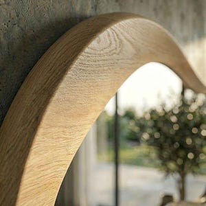 May include: A curved mirror frame crafted from light-toned natural wood with a visible grain pattern. The frame features a smooth, rounded edge and a thick, solid construction that follows a gentle arch, reflecting a blurred outdoor scene.
