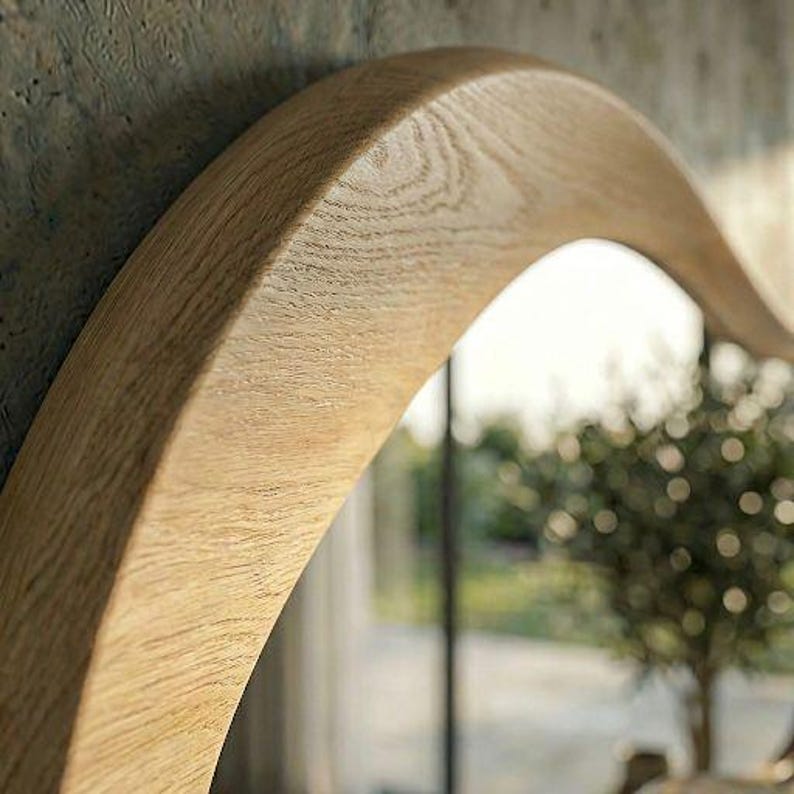 May include: A curved mirror frame crafted from light-toned natural wood with a visible grain pattern. The frame features a smooth, rounded edge and a thick, solid construction that follows a gentle arch, reflecting a blurred outdoor scene.