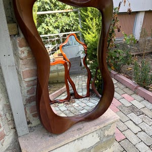 May include: A wall mirror featuring a thick, polished dark brown wooden frame with an organic, asymmetrical, and wavy silhouette. The reflective glass surface captures a clear view of the surrounding outdoor environment and a smaller mirror.