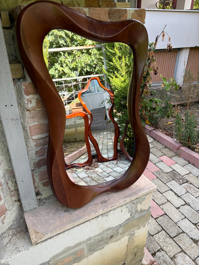 May include: A wall mirror featuring a thick, polished dark brown wooden frame with an organic, asymmetrical, and wavy silhouette. The reflective glass surface captures a clear view of the surrounding outdoor environment and a smaller mirror.
