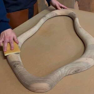 May include: An unfinished, light-colored wooden frame with an irregular, organic, wavy shape. The wood displays visible natural grain patterns and light brown tones. A rectangular sanding block is being used to smooth the surface of the frame.