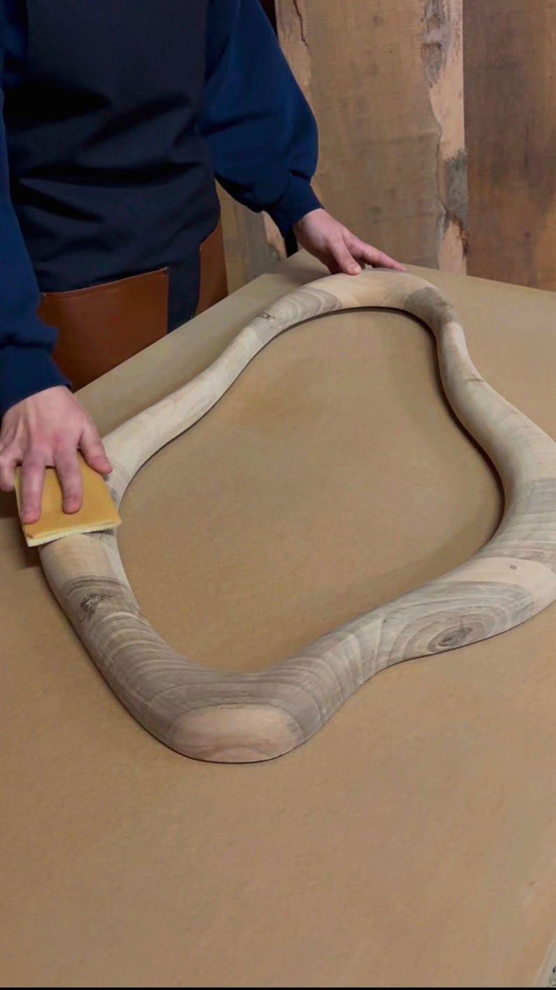May include: An unfinished, light-colored wooden frame with an irregular, organic, wavy shape. The wood displays visible natural grain patterns and light brown tones. A rectangular sanding block is being used to smooth the surface of the frame.