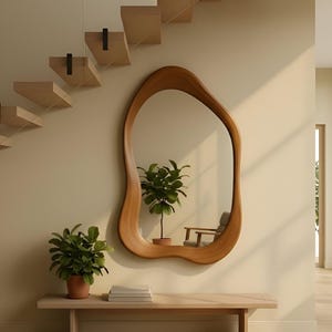Puede incluir: Un espejo de pared con forma org&aacute;nica irregular y un marco de madera lisa de color marr&oacute;n claro. El espejo est&aacute; montado en una pared beige sobre un banco de madera clara, que sostiene una planta verde en maceta y libros.