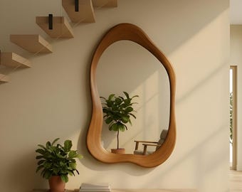 May include: An irregular, organic-shaped wall mirror with a smooth, light-brown wooden frame. The mirror is mounted on a beige wall above a light-coloured wooden bench, which holds a potted green plant and a small stack of books.