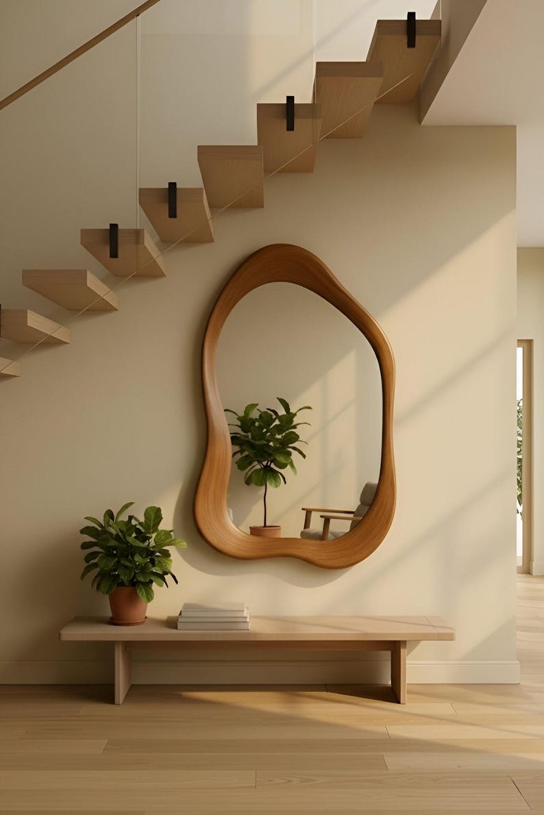 Puede incluir: Un espejo de pared con forma org&aacute;nica irregular y un marco de madera lisa de color marr&oacute;n claro. El espejo est&aacute; montado en una pared beige sobre un banco de madera clara, que sostiene una planta verde en maceta y libros.