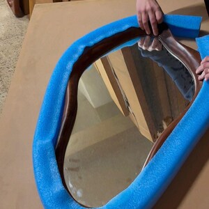 May include: An irregular, organic-shaped mirror with a dark brown wooden frame. The mirror is partially wrapped in bright blue foam protective padding, which contours around the curved edges of the wooden frame to provide cushioning for the glass.