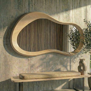 May include: A wall-mounted mirror with an irregular, organic-shaped light wood frame. The reflective surface shows a reflection of a wooden wall paneling and a green plant. The mirror is positioned above a long, rectangular bench with a tan cushion.
