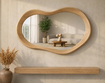 May include: An irregular, organic-shaped wall mirror with a light-toned wooden frame. The mirror reflects a living room interior featuring a beige sofa, a small wooden coffee table, and a potted plant. Below the mirror is a long, rectangular wooden shelf.