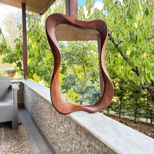 May include: A wall mirror featuring a smooth, dark brown wooden frame with a distinctively fluid, organic, and undulating shape. The reflective glass surface is encased within the curved, polished wood border, creating a continuous, flowing silhouette.