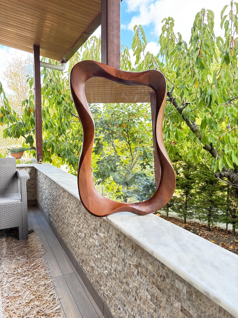 May include: A wall mirror featuring a smooth, dark brown wooden frame with a distinctively fluid, organic, and undulating shape. The reflective glass surface is encased within the curved, polished wood border, creating a continuous, flowing silhouette.