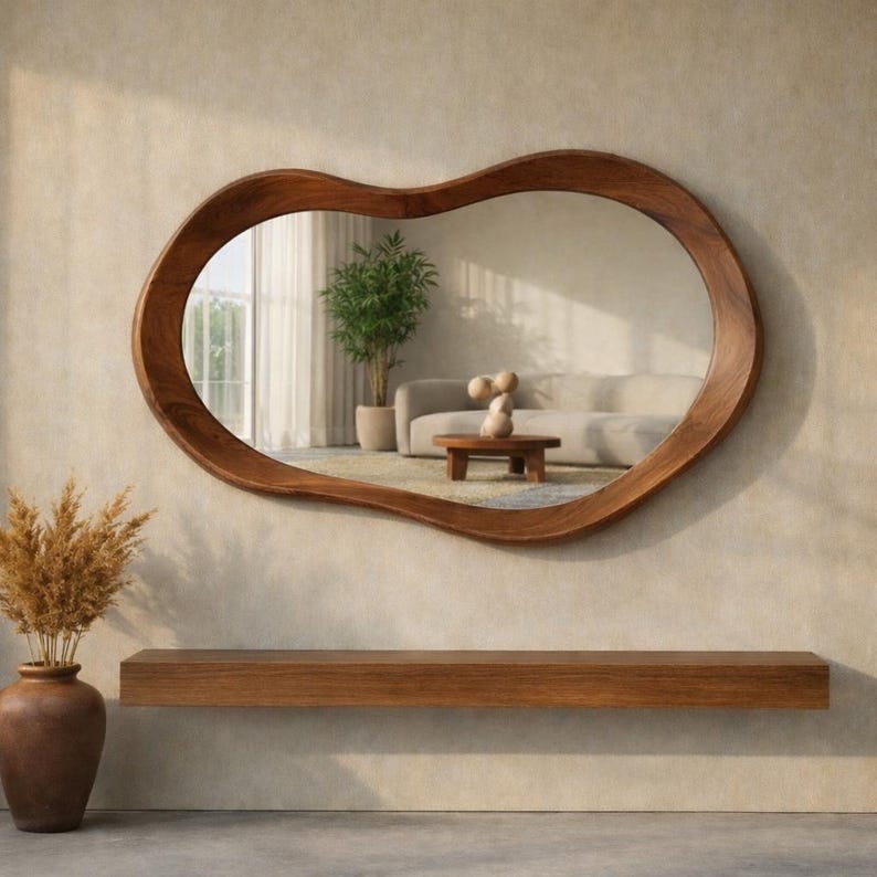 May include: A wall-mounted mirror with an irregular, organic-shaped wooden frame. The mirror reflects a living room interior featuring a light-colored sofa, a small wooden coffee table, and a potted plant. Below the mirror is a wooden shelf.
