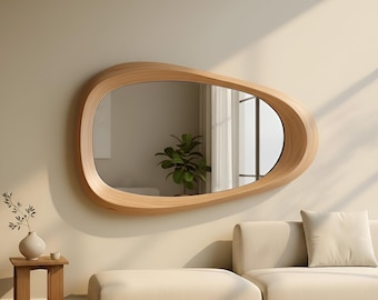 May include: A large, modern mirror with a unique, organic shape. The mirror is framed in light-colored wood and hangs on a neutral-toned wall. The reflection shows a glimpse of a room with a plant and a window. The overall aesthetic is minimalist and contemporary.
