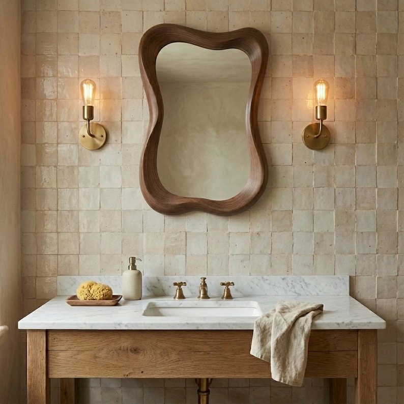 May include: A bathroom vanity featuring a white marble countertop, a wooden cabinet base, and a wavy-edged wooden mirror. Two brass wall sconces with exposed bulbs flank the mirror, and a beige towel rests on the counter near a soap dispenser.