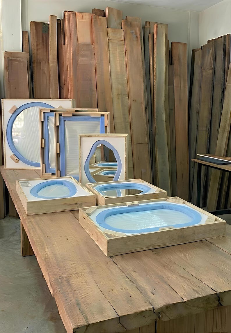 May include: Several mirrors with light blue foam protective borders are secured inside light-colored wooden shipping crates. The mirrors vary in shape, including circular and oval designs, and are arranged on a large wooden workbench.