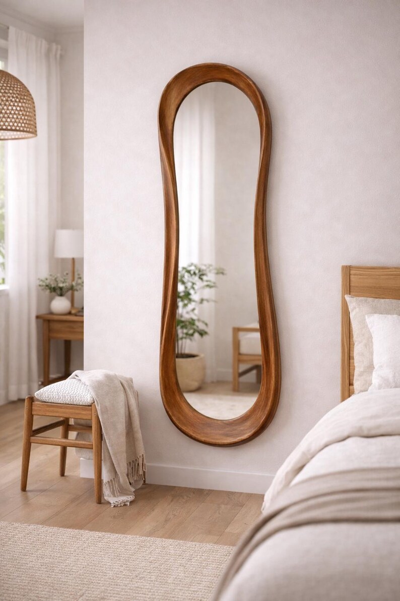 May include: A full-length wall mirror featuring a thick, organic, wavy wooden frame with a warm brown finish. The mirror has an elongated, irregular oval shape that tapers and curves along the sides, creating a fluid, non-symmetrical silhouette.