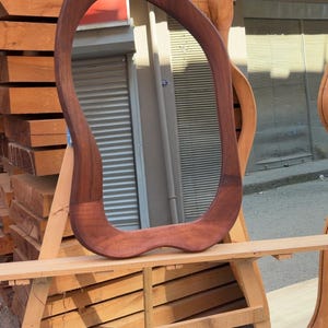 Puede incluir: Un espejo de pared con un marco de madera de color marr&oacute;n oscuro de forma irregular y org&aacute;nica. El espejo presenta una silueta curva y suave, con una superficie de cristal reflectante que muestra una vista distorsionada del entorno.