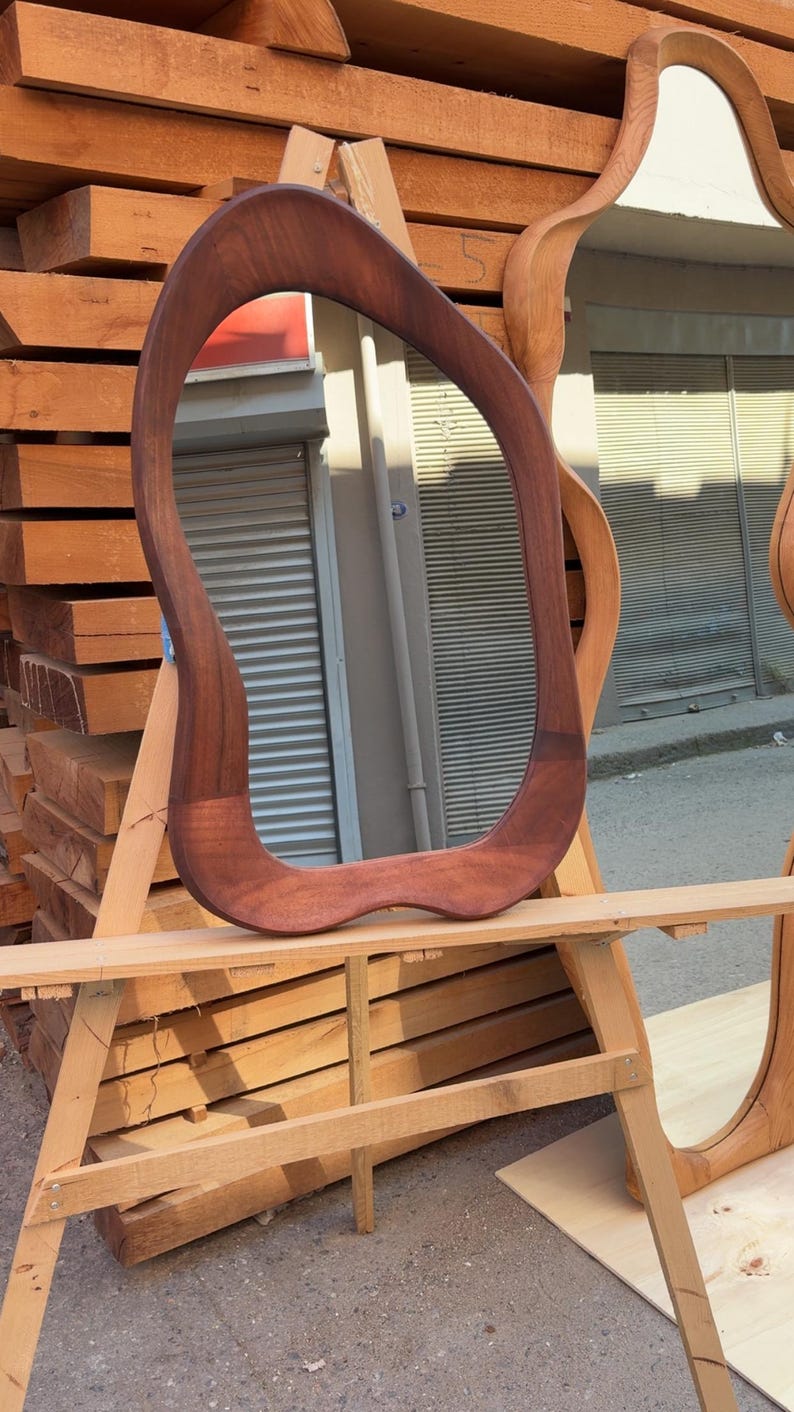 Puede incluir: Un espejo de pared con un marco de madera de color marr&oacute;n oscuro de forma irregular y org&aacute;nica. El espejo presenta una silueta curva y suave, con una superficie de cristal reflectante que muestra una vista distorsionada del entorno.