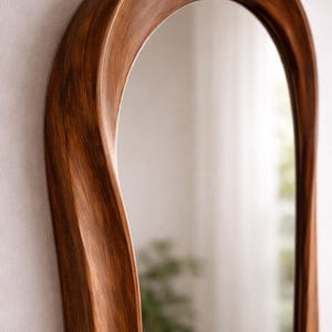 May include: A wall mirror featuring a tall, arched frame crafted from dark brown wood with a visible grain pattern. The frame has a smooth, undulating, and curved silhouette that creates a fluid, organic shape around the reflective glass surface.