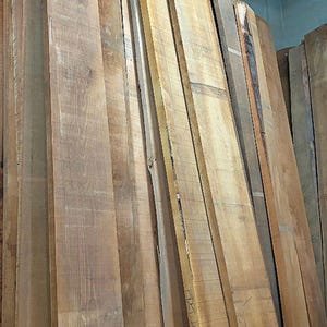 May include: A stack of long, unfinished wooden planks standing vertically. The boards feature natural light to medium brown wood grain patterns, with some edges showing raw bark. One board has the handwritten text ADET-A1 visible on its surface.