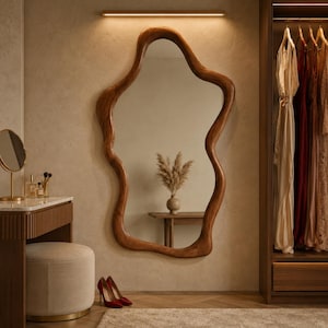 May include: A large, irregular, organic-shaped wall mirror with a thick, polished wooden frame. The mirror is mounted on a textured beige wall, positioned above a wooden console table, reflecting a vase with dried pampas grass and a wooden table.