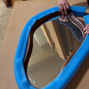 May include: An irregular, organic-shaped mirror with a dark brown wooden frame. The mirror is partially wrapped in bright blue foam protective padding, which contours around the curved edges of the wooden frame to provide cushioning.